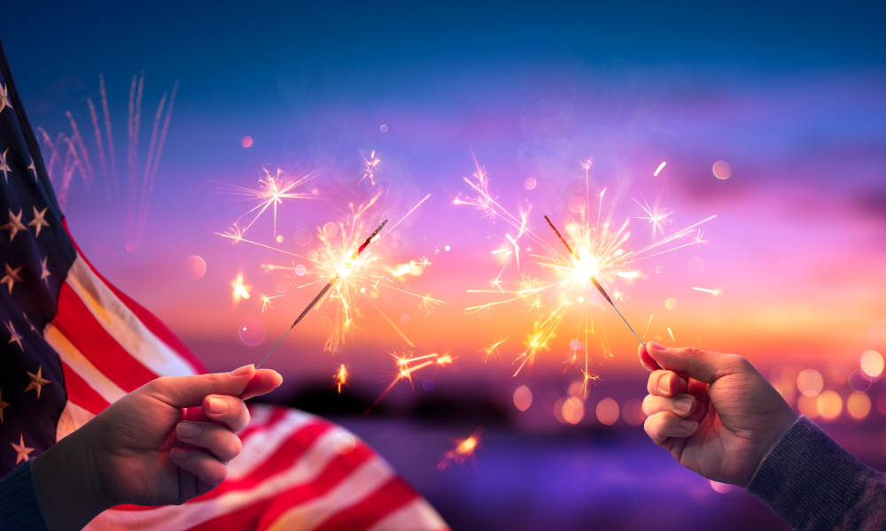 Two hands holding sparkler fireworks with a background of an American flag at sunset.