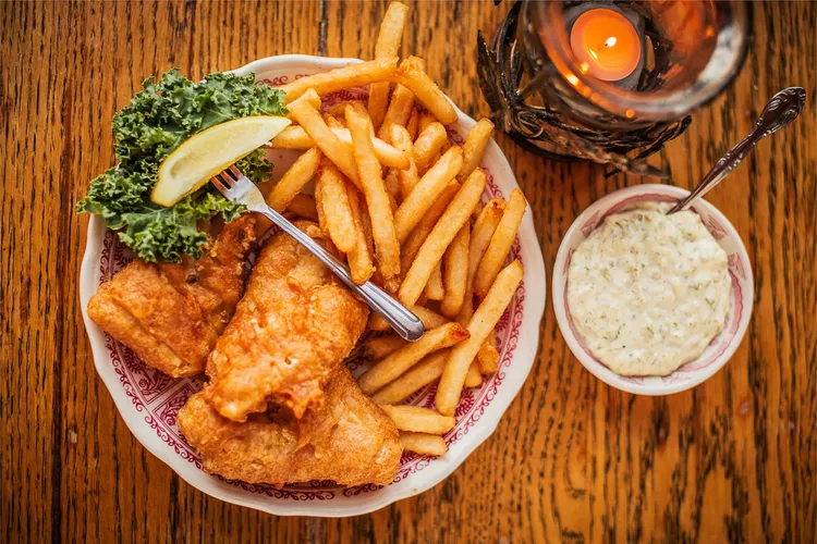 fried fish, French fires, kale, a lemon wedge on a fork, a bowl with sauce and a spoon, and a candle in a holder