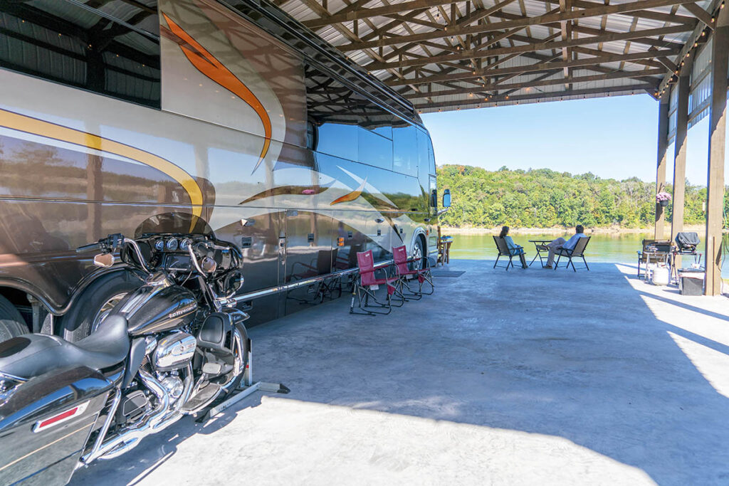 RV beside a motorcycle on a concrete pad with an overhead cover on the lake shore.