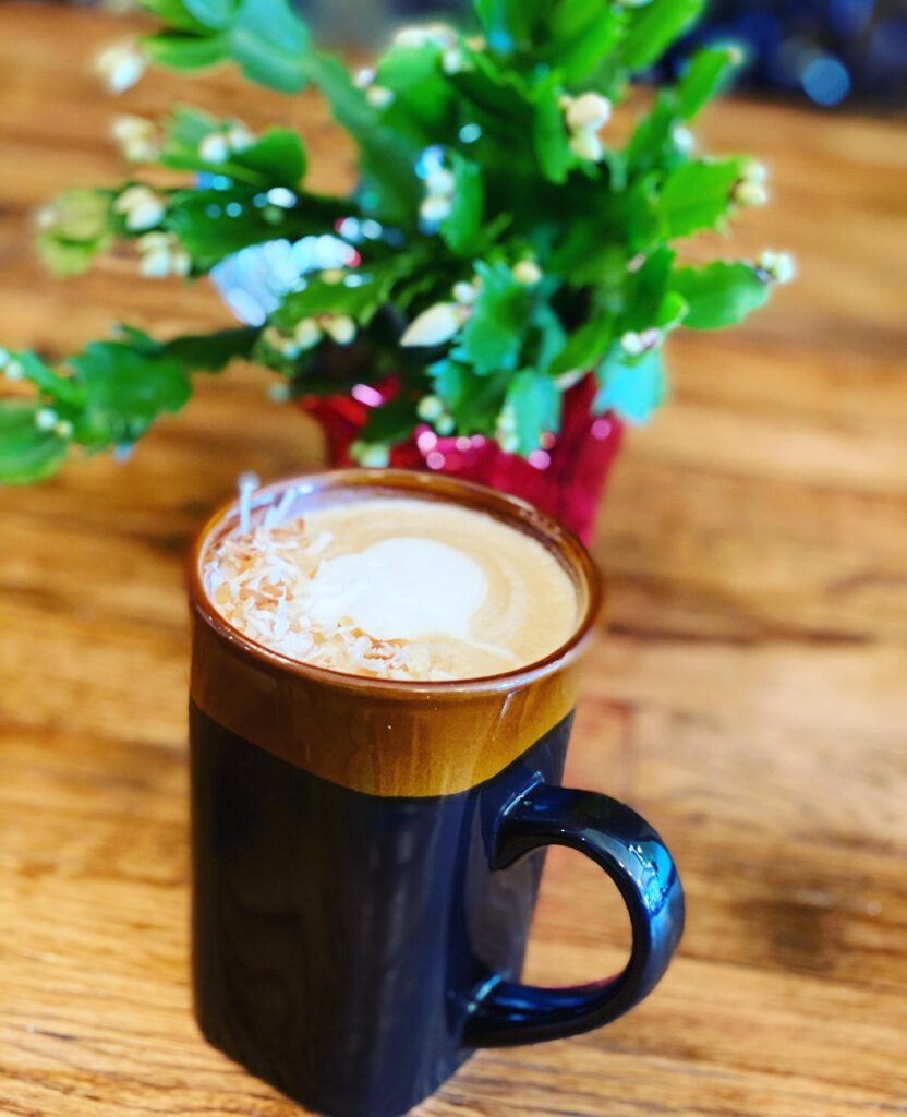 A latte with coconut shavings in front of a green plant with a red pot. 
