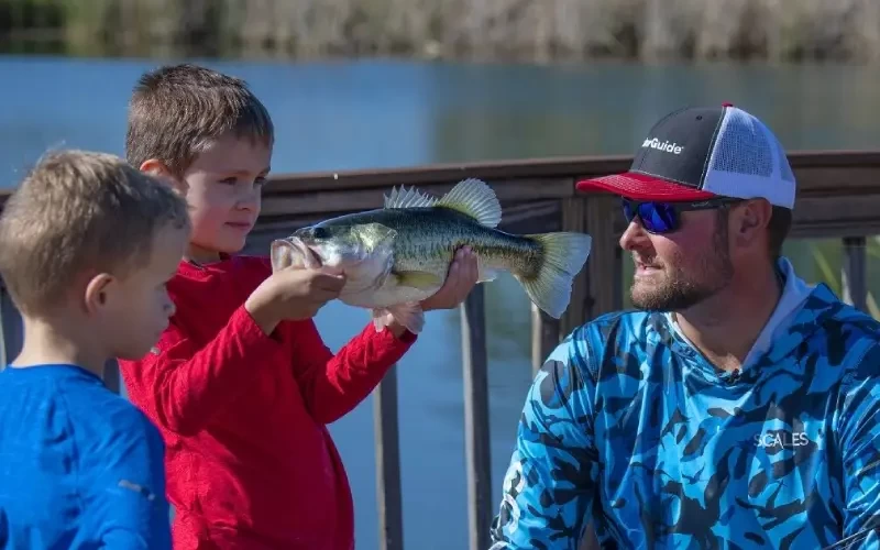 Boy holding fish beside other boy while a man with a hat and sunglasses looks on.