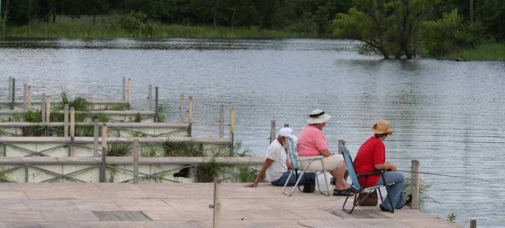 Two women in lawn chairs sitting beside a man on a dock while fishing on the lake.