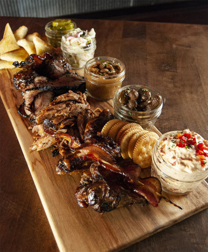 A Southern BBQ board with peanuts, pickles, slaw, crackers, bread, and sauce on a wooden table. 