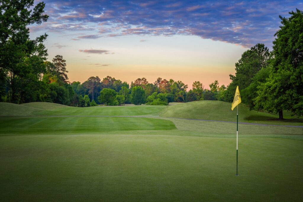Golf course featuring a yellow flag over a hole with rolling hills and sunset in the background.